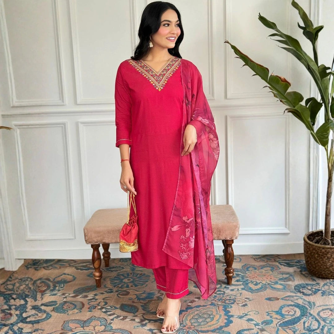 Woman in a red traditional outfit standing in a room with a plant and bench.